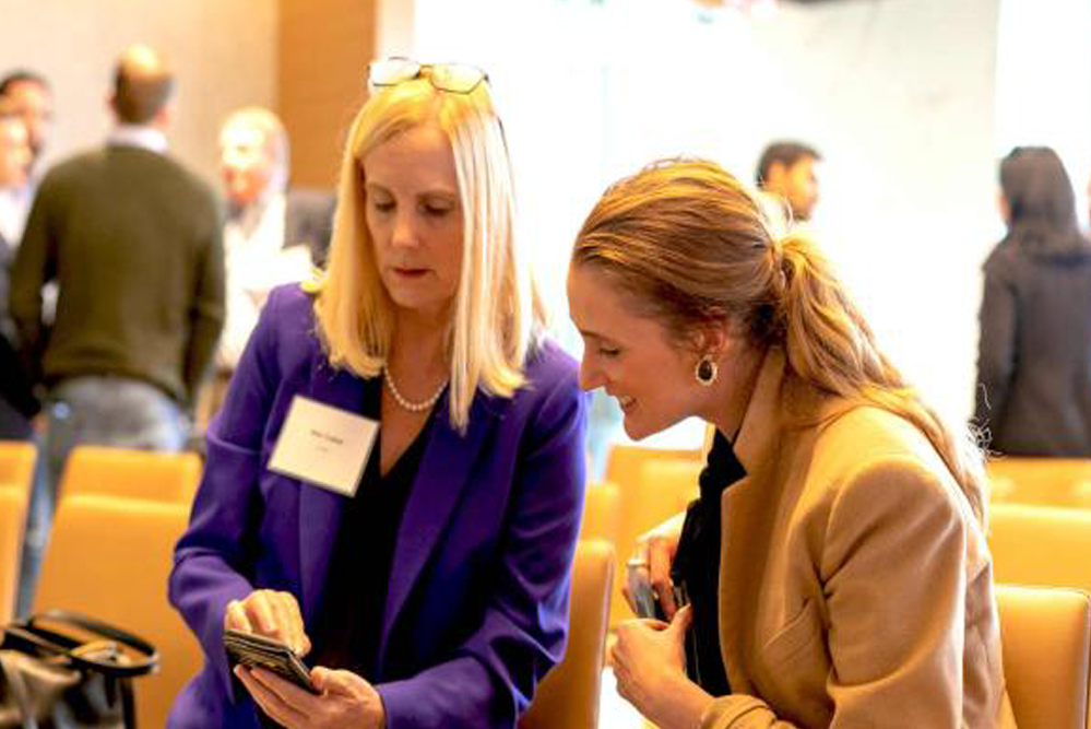 Two attendees seated in an auditorium review information on a smartphone during a networking event, with other participants and rows of chairs visible in the background.