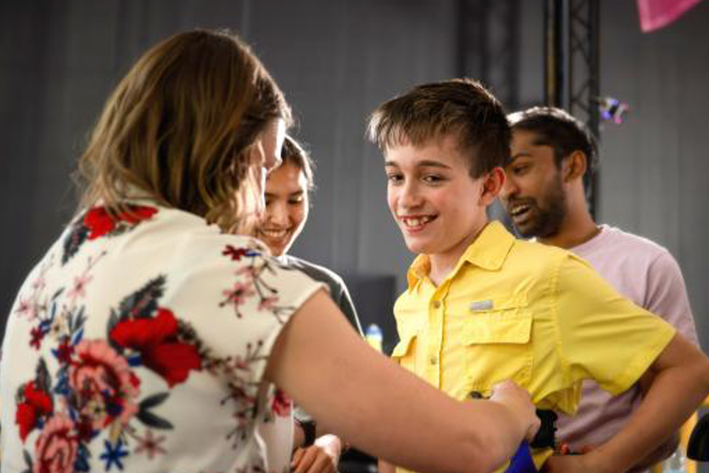 An adult places a ribbon around a child’s neck during an onstage event, with another child standing nearby and event equipment visible in the background.