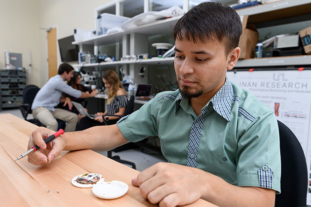 two males working in a lab, one in foreground sitting at table while examining a round white device