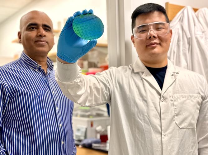 Two researchers in a laboratory examine a blue-green silicon wafer. One, wearing a lab coat, safety goggles, and a blue glove, holds the wafer up for inspection while the other stands beside them. Laboratory equipment is visible in the background.